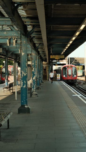 A view of East Croydon Station platform showing an overhead metal canopy supported by weathered blue-painted steel columns. The platform surface is paved with grey concrete tiles, featuring tactile paving strips near the train tracks for accessibility. On the right, a red electric train is stationary at the platform, with its doors closed, ready for passenger boarding or disembarking. A person dressed in casual clothing is walking along the platform towards the train, carrying a small bag. To the left, there are a few benches and some electrical and signaling equipment mounted on the platform. The station is illuminated by natural daylight, with a partially visible background showing additional tracks and station structures. This setting is associated with the logistics of home relocation and furniture transport, as part of house removals and moving services facilitated by Man and Van Croydon.