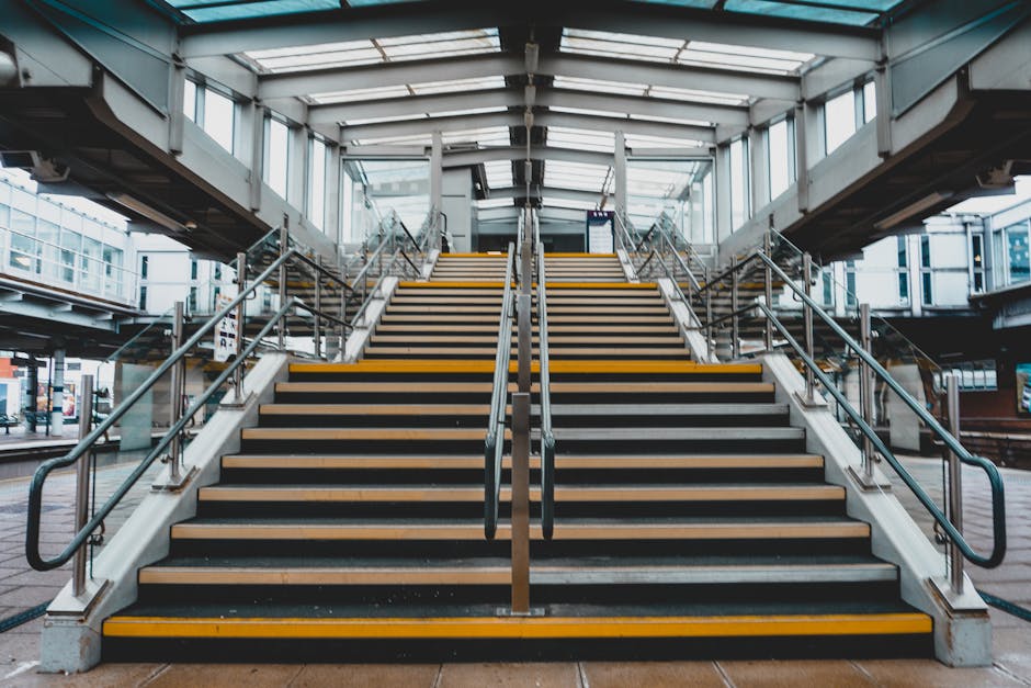 A wide staircase leading up to the entrance of East Croydon Station, with multiple steps covered in black anti-slip material and highlighted with yellow safety strips along the edges. Silver metal handrails run parallel on both sides and down the center of the stairs, providing support for users during the loading process. The stairway is situated indoors within the station, under a clear glass roof structure that allows natural light to illuminate the area. To the sides and behind the stairs, glass barriers and metal framing are visible, opening onto the platform area. The station environment appears clean and modern, with a focus on accessibility for commuters and movers. The scene suggests an active setting for home relocation or furniture transport, where removal companies like Man and Van Croydon might assist in moving household items through efficient stairs and loading procedures.