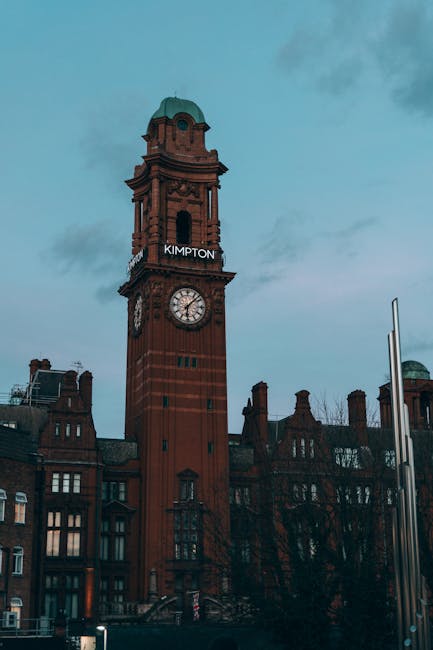 A tall, historic brick clock tower with ornate architectural details, including arched windows and a decorative balcony near the top, is shown in the image. The clock face is clearly visible, displayi