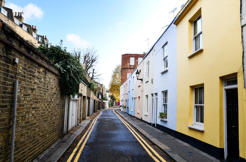 A narrow residential street in South Croydon with a slight curve, featuring a mix of traditional brick houses on the left and colorful, modern-facing terraced houses on the right, including yellow and white façades. The street is paved with asphalt and has double yellow lines along the curbstones, indicating parking restrictions. The pavement on both sides is made of concrete slabs, and some houses display small front gardens with potted plants. The scene is captured during daylight with a partly cloudy sky, and a few trees are visible in the background, some with autumn foliage. The street appears to be quiet, suitable for household furniture transport and home relocation activities, with no visible vehicles or moving equipment present. Occasionally, [COMPANY_NAME]'s house removals services may utilise such streets for efficient loading and unloading during home moves, especially where access is constrained by narrow street layouts.