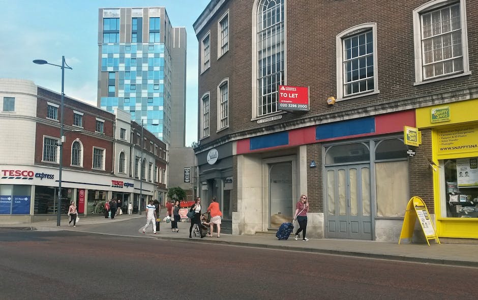 Street view in Croydon showing pedestrians walking along the pavement in front of a row of mixed-use buildings, including a Tesco Express store on the left with signage and shop windows. To the right, a large brick building features multiple tall, arched and rectangular windows, and a red and blue awning above boarded-up entrances. A woman with sunglasses and a pink top is pulling a blue suitcase along the sidewalk, while other pedestrians are crossing or walking along the street. In the background, tall modern office or apartment buildings with reflective glass facades rise above the older, lower-rise structures. The scene is captured in daylight with a clear sky, and the environment suggests a busy urban area suitable for house or shop relocations. Man and Van Croydon occasionally services such areas, supporting packing and furniture transport during home relocation or commercial moves.