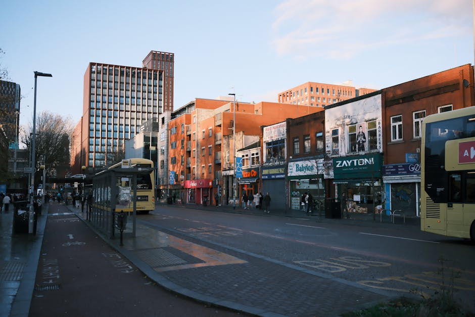 A wide view of a busy urban street scene during daytime features several multi-storey buildings with a mix of modern and older architectural styles. The buildings house various shops with storefront signage, including a pizza restaurant, a baby shop, and other retail outlets, with some featuring large billboard advertisements on their facades. The street is populated with pedestrians walking along the pavement. Two double-decker buses are parked at bus stops on either side of the street, with one bus partially visible on the right and the other further down the road. The road layout includes designated bus lanes marked with yellow paint, and the pavement has tactile paving for visually impaired pedestrians. Several streetlights, trees, and lampposts are visible along the sidewalk. The weather appears clear, with bright daylight illuminating the scene, suggesting a typical day for local retail activity and public transport, relevant to house and business relocations managed by Man and Van Croydon, as seen on the page titled Waddon to Purley Way commercial removals for shops.