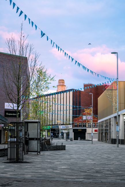 Street view in Croydon showing pedestrians walking along the pavement in front of a row of mixed-use buildings, including a Tesco Express store on the left with signage and shop windows. To the right, a large brick building features multiple tall, arched and rectangular windows, and a red and blue awning above boarded-up entrances. A woman with sunglasses and a pink top is pulling a blue suitcase along the sidewalk, while other pedestrians are crossing or walking along the street. In the background, tall modern office or apartment buildings with reflective glass facades rise above the older, lower-rise structures. The scene is captured in daylight with a clear sky, and the environment suggests a busy urban area suitable for house or shop relocations. Man and Van Croydon occasionally services such areas, supporting packing and furniture transport during home relocation or commercial moves.